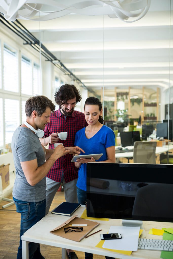 Three professionals at Shivah Web Tech stand in an office, examining a tablet together with focused expressions.